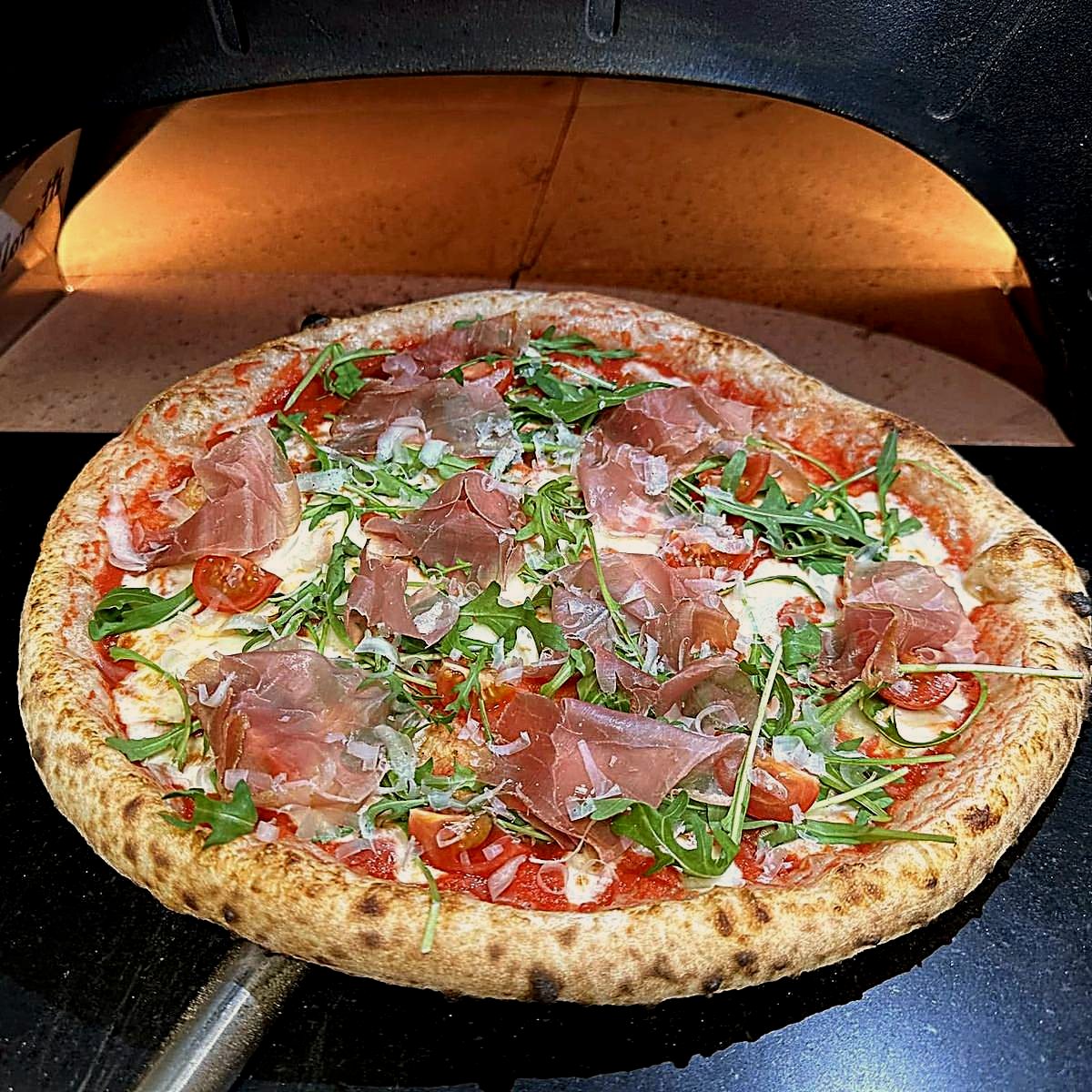 Egész pizza a kemence izzó szája előtt, prosciutto crudo szeletekkel, rukkolával és koktélparadicsommal megrakva
