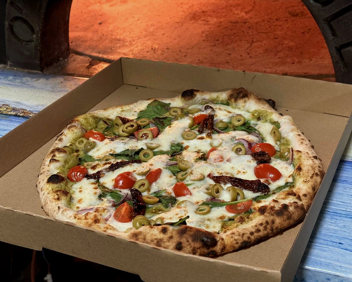 Nyitott pizzásdobozban álló, egész frissen sült pizza, a háttérben a kemence izzó boltíve