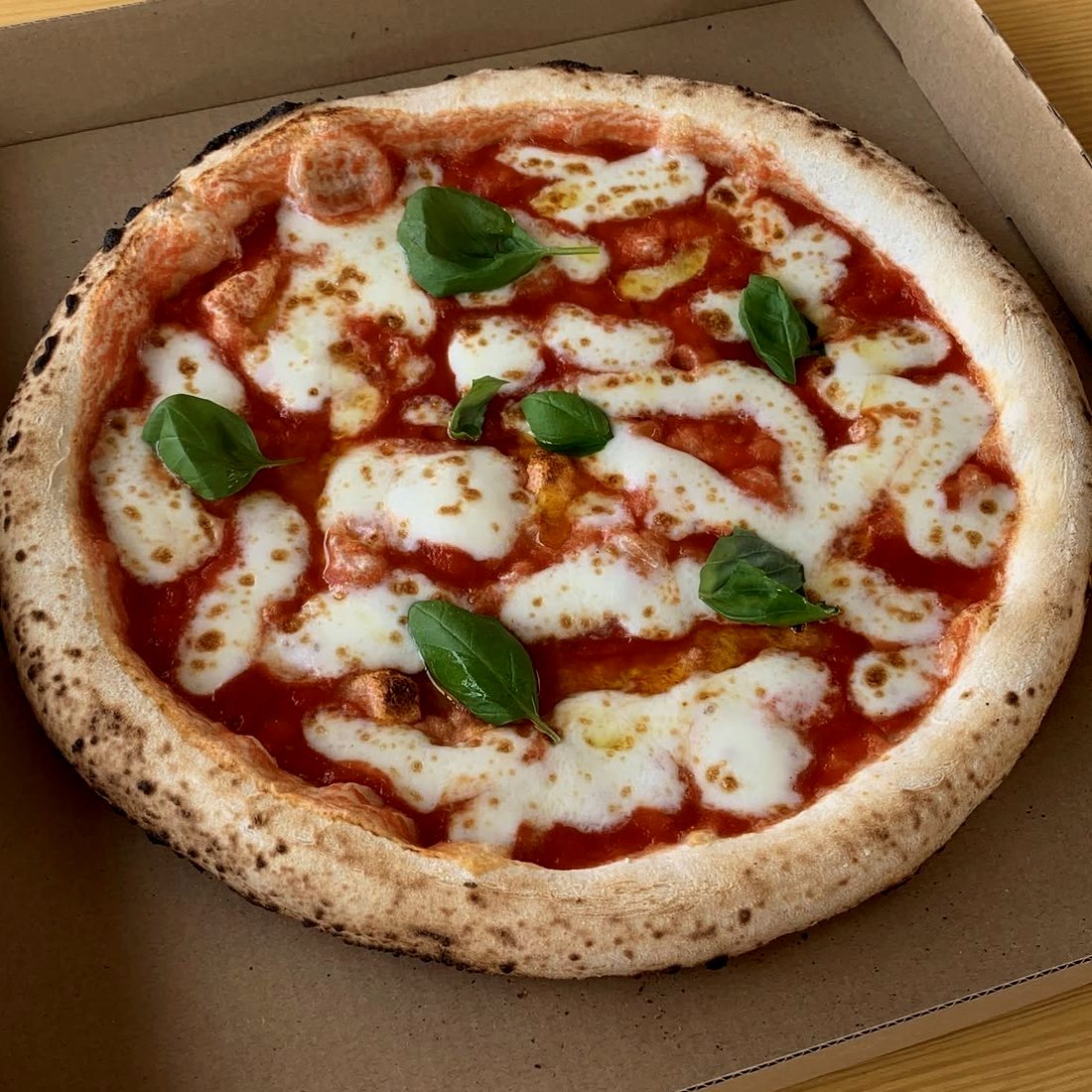 Egész margherita pizza nyitott dobozban, felülnézetből: paradicsom-alap, olvadt mozzarella-foltok és friss bazsalikom