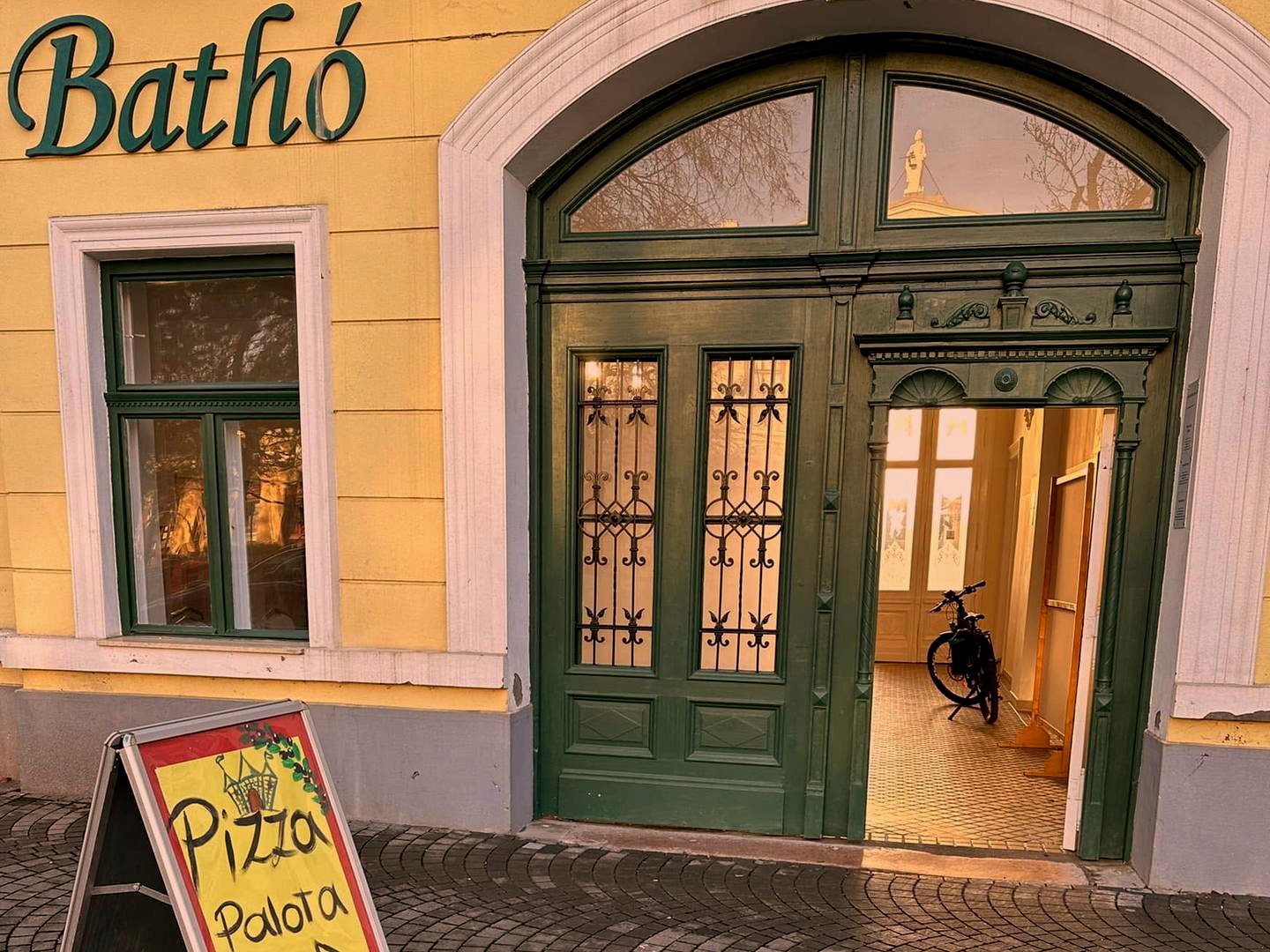 A Bathó Palota sárga homlokzata a Lehel vezér téren, zöld kapuval és a Bathó felirattal, előtte a Pizza Palota kihelyezett táblája
