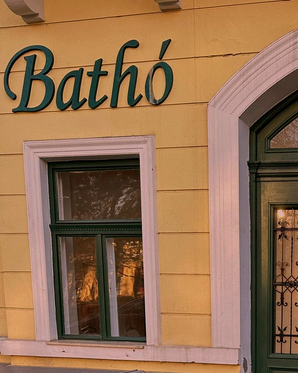 A Bathó felirat a palota sárga homlokzatán, mellette zöld keretes ablak és a boltíves kapu kőkeretének indulása