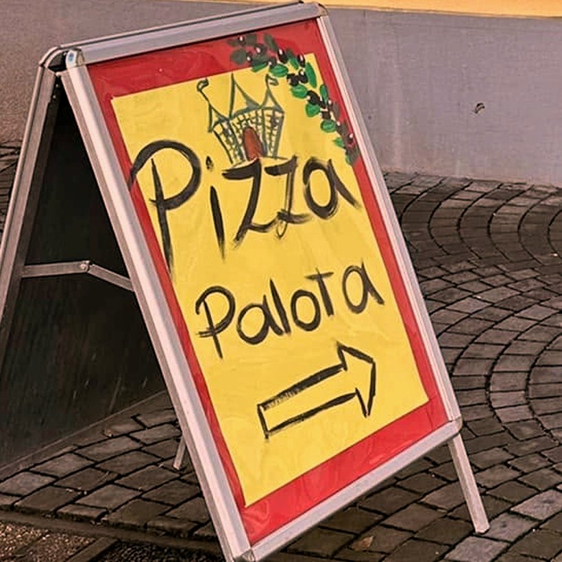 A pizzéria kézzel festett, sárga kihelyezett táblája a Pizza Palota felirattal és nyíllal a kapualj felé