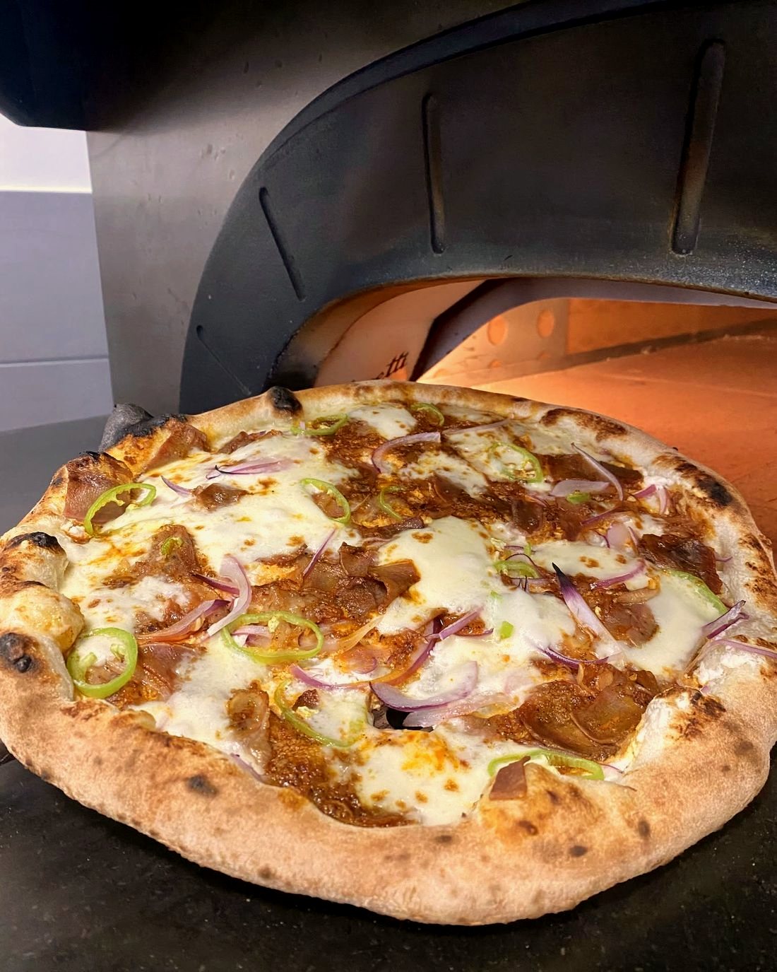 Egész, frissen sült pizza a kemence izzó szája előtt, lilahagymával, zöld hegyes paprikával és füstölt hússal megrakva